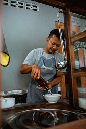 man seller in apron pouring vegetable oil from a bottle to bowl when preparing chicken noodles dishの写真素材