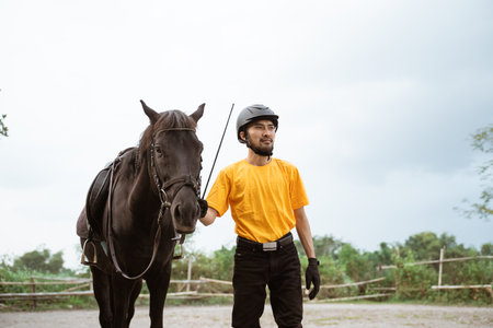 Male equestrian in equipment standing next to horse in field arenaの写真素材