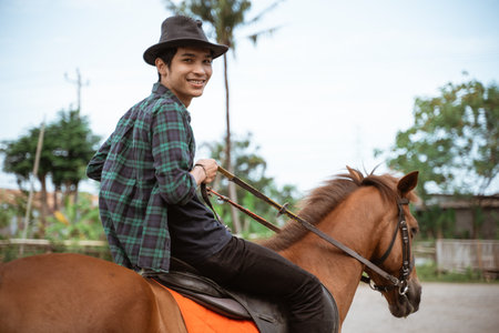young man wearing cowboy hat smiling while going horseback riding on outdoor backgroundの写真素材