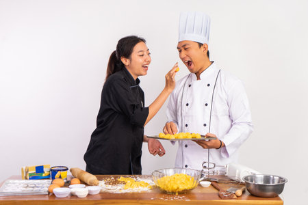 female chef feeding male chef a cake while carrying a baking sheet on isolated backgroundの写真素材