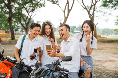 Four high school students laugh after looking at cellphone screen while sitting on motorbikeの写真素材