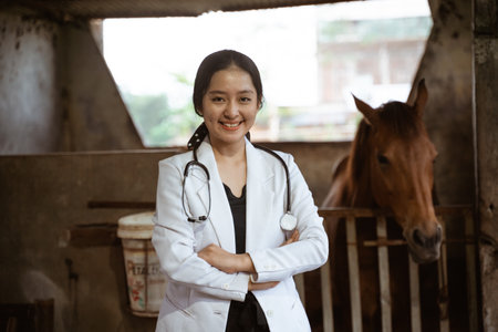 smiling asian female veterinarian standing with horse stable in the backgroundの写真素材
