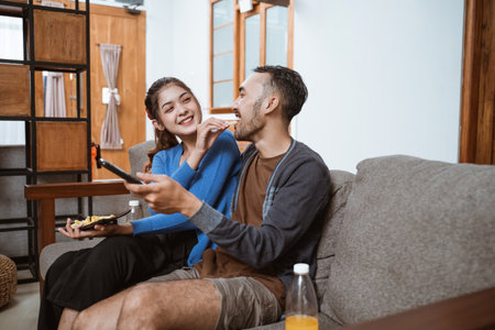woman feeding her boyfriend a snack sitting on the couch while watching together at homeの写真素材