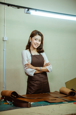 asian craftwoman wearing apron standing with crossed hands in tanned workspace