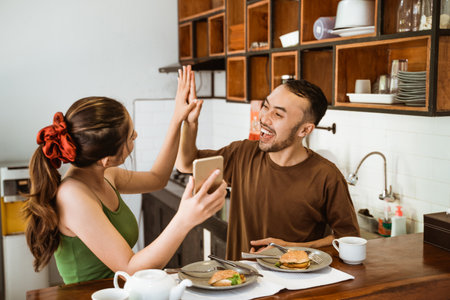 asian couple with happy applause while looking at smartphone screen while having breakfast in dining roomの写真素材