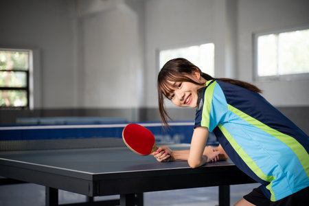 female athlete smiling while leaning on table tennis table and holding paddle in indoor buildingの写真素材
