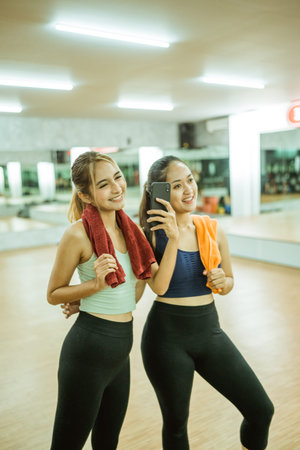 two attractive women holding towels during break and photo using smartphone camera in fitness center...