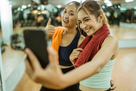 two happy asian women taking selfies together using a cellphone camera after a workout at the gymの写真素材
