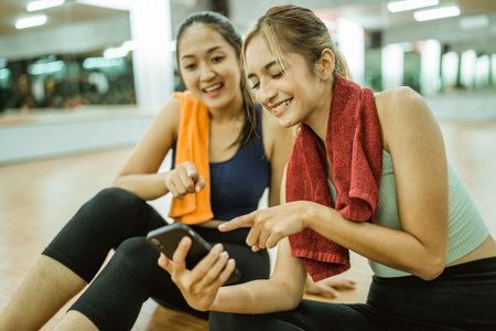two asian women sitting while using mobile phone post workout at gym