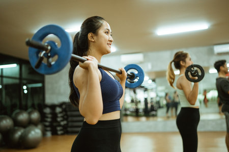 young asian woman lifting barbell in front of chest during exercise in fitness roomの写真素材