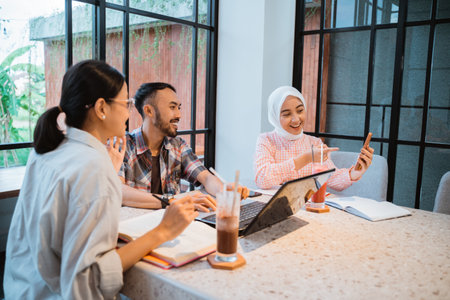three smiling college students looking at mobile phone screens together during group work in a cafeの写真素材