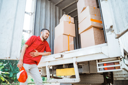 Delivery man having stomach ache while working with cardboard boxes behind container truck as...