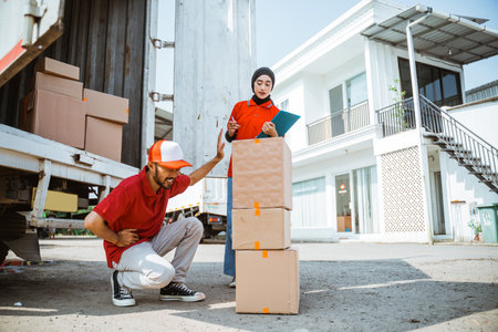 delivery man in red uniform squatting with stomach ache while lifting box next to female worker standing behind container truckの写真素材