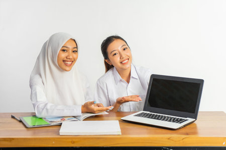 two high school girls sitting at desk with tablet and books while showing laptop screen on isolated...