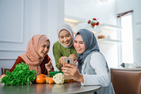 happy young asian woman with hijab cooking in the kitchen while looking at internet on mobile phone for menu todayの写真素材