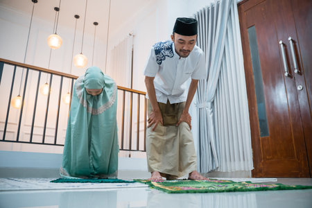 Asian husband leads prayer with wife during Ruku movement when praying on a prayer mat at homeの写真素材