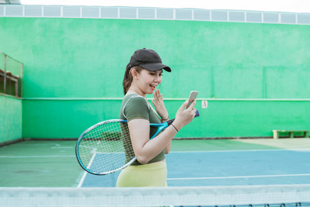 Female tennis player holding racket shocked while looking at mobile phone screen on tennis courtの写真素材