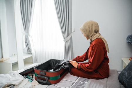 Asian woman in veil puts clothes in a suitcase while sitting on the bedの写真素材