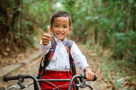 a male elementary student in uniform riding his bike through the country road with thumb up and a...