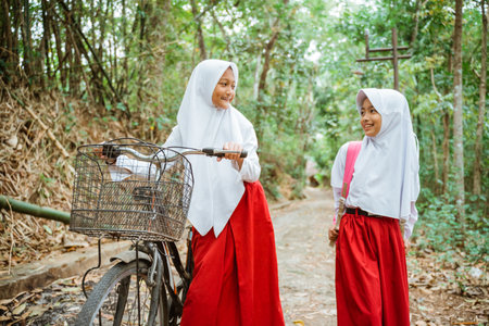two female elementary students in uniform walking together through the country road and smiling with a lot of tree at the backgroundの写真素材