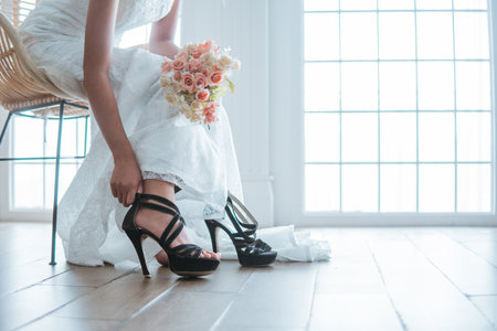 asian bride in white gown wearing the black heels while holding the flowers bouquet and sitting on the chairの写真素材