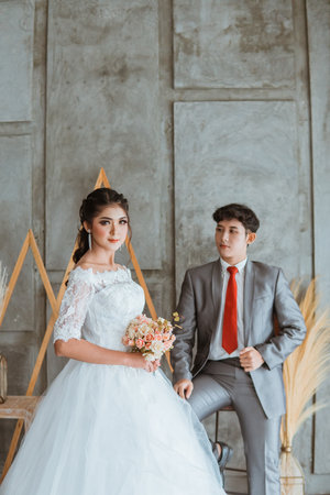 asian groom touch his suit while sitting on the chair behind the beautiful bride in white gown that holding the flowers bouquetの写真素材