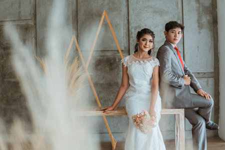 beautiful asian bride in white gown holding the flowers bouquet while sitting beside the groom at photo studioの写真素材