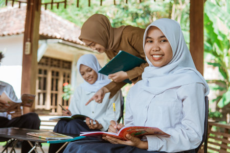Middle school girls wearing headscarves smiling holding pens and books sitting in the gazebo during outdoor classの写真素材
