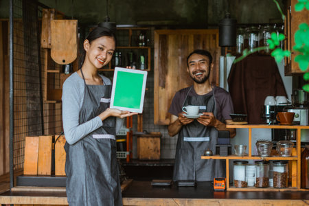 female barista in apron standing showing the digital tablet with green screen with the male barista standing at the background holding a cup of coffeeの写真素材