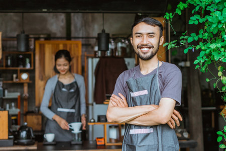 male barista in apron standing with handcrossed and smiling with the female barista brewing the coffee at the backgroundの写真素材