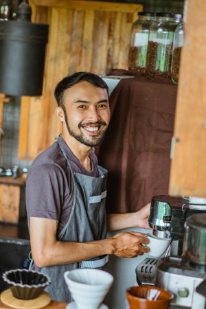asian male barista in apron standing with smile while operating the coffee machine at the coffee shopの写真素材