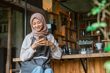 female barista with hijab and apron using the phone while sitting in front of the bar deskの写真素材