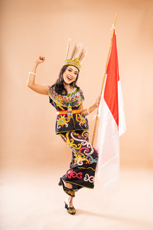 asian woman in traditional clothes of dayak tribe standing with clenched arm and bend her leg while holding the indonesian flag on isolated backgroundの写真素材