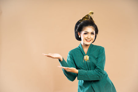 asian woman in green kebaya standing and pointing on her side with her both hands on isolated backgroundの写真素材