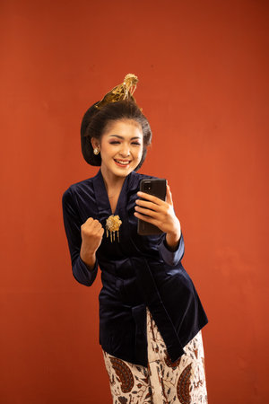 javanese woman in bludru kebaya clenched her hand while looking at the phone on isolated abckgroundの写真素材