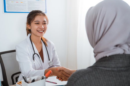 a beautiful doctor in doctors coat with the stethoscope on her shoulder hand shake with the female patient in grey veilの写真素材