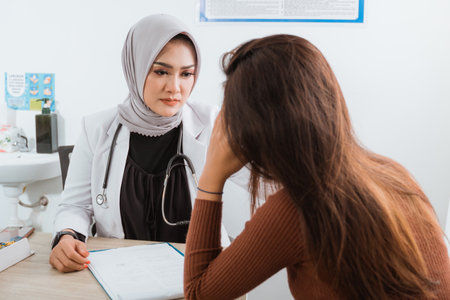 a female doctor in doctors coat with the stethoscope at her shoulder look at the female patient that touch her head in painの写真素材