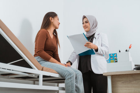 the female patient sitting on the bed while the female doctor in doctors coat standing next to her with the blue clipboard at her handの写真素材