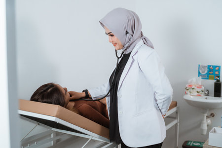 a beautiful doctor in doctors coat smiling while checking the female patient that laying on the bed using the stethoscopeの写真素材