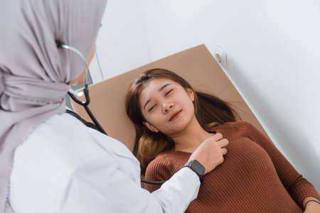 a beautiful patient laying on the bed and close her eyes while checked by the female doctor using the stethoscopeの写真素材