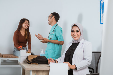 a female doctor in doctors coat sitting on her desk and smiling while the male nurse talking to the female patient at the backgroundの写真素材