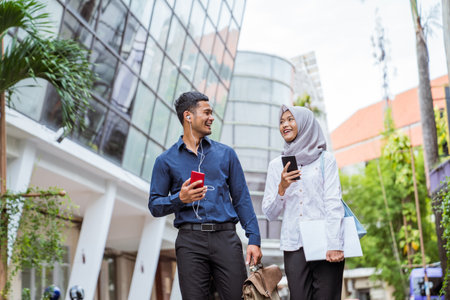 Asian male and female Muslim couple chatting and holding cellphones while walking to work togetherの写真素材