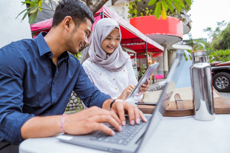Asian female worker in hijab and male worker using digital tablet and laptop sitting in outdoor...