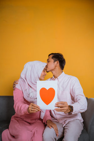 Asian man kisses hijab woman on the forehead holding and showing a paper love drawing while sitting on a sofaの写真素材