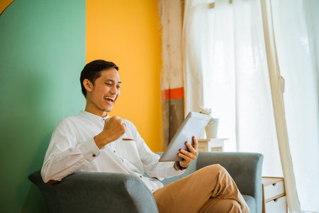 portrait of excited young Asian man sitting on couch using pad at homeの写真素材