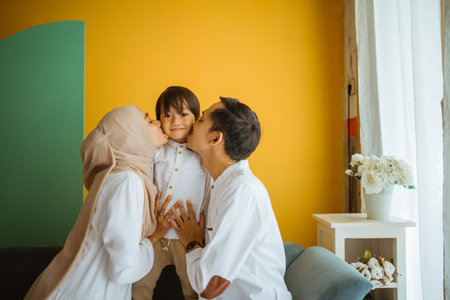 Asian Muslim mom dad embrace and kiss son sitting on sofa in living roomの写真素材