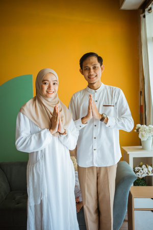 young Asian couple standing with greeting hands gesture during Eid in the living room