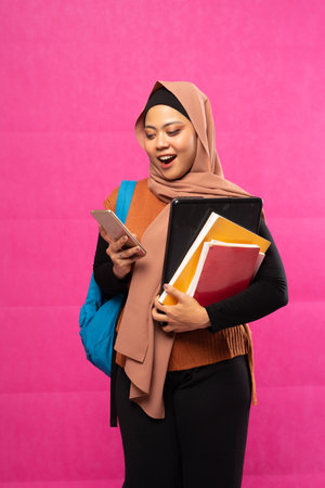 shocked Asian Muslim student stands with bag and book looking at cell phone in isolated backgroundの写真素材
