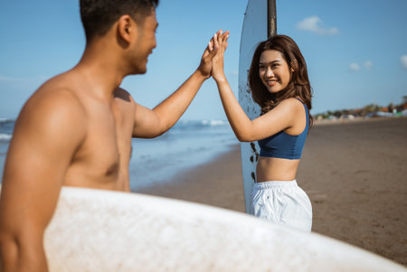 couple holding a surfboard and giving a high five to each other on a blue sky beachの写真素材
