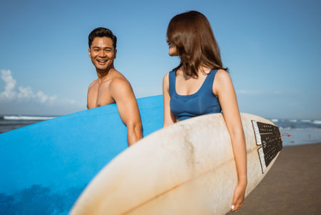 Asian couple carrying surfboards chatting at beach against blue sky enjoying a vacationの写真素材